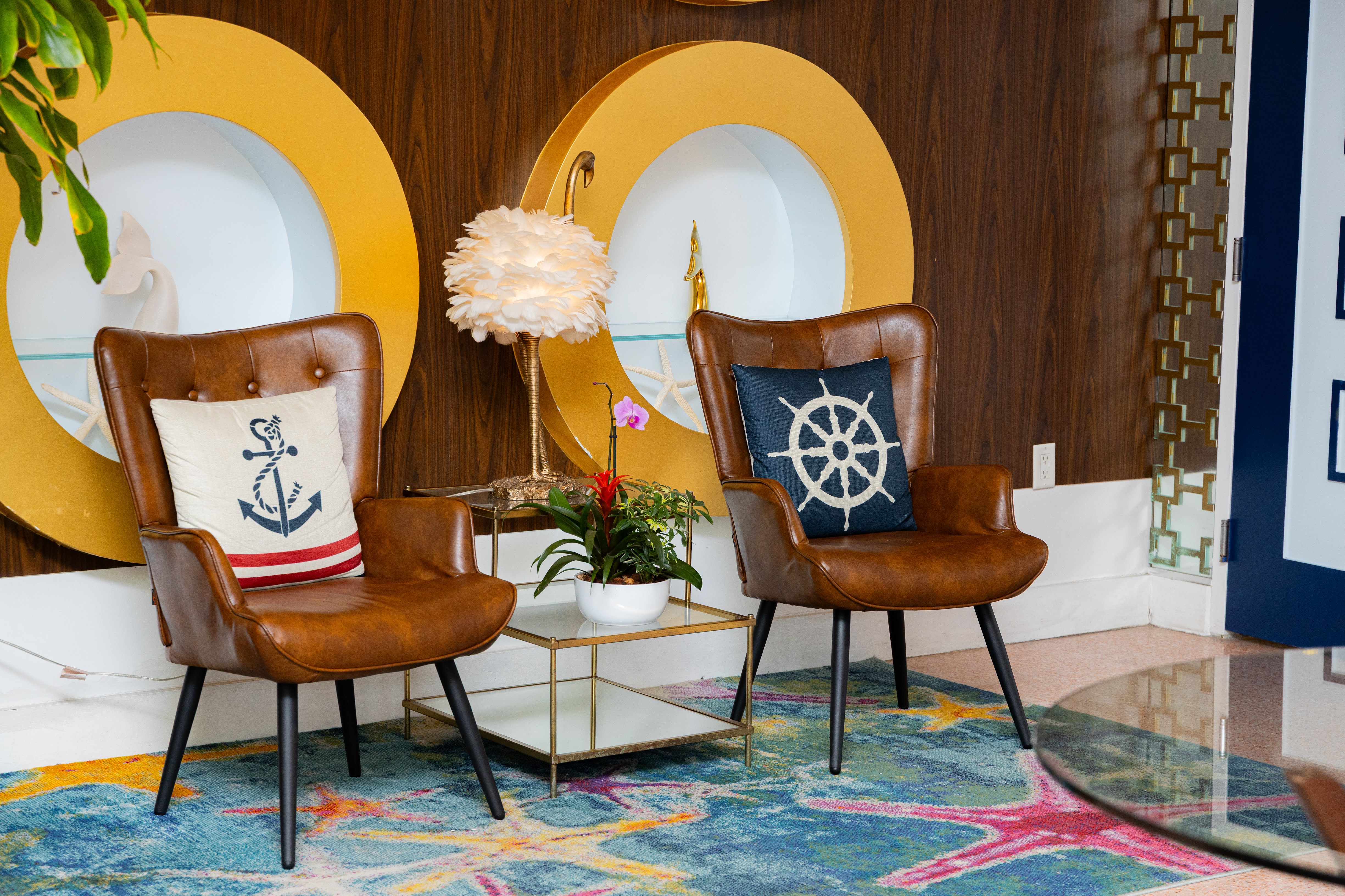 Two leather chairs with nautical-themed pillows sit in a lobby area with a colorful rug and a coffee table.