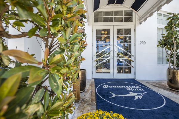 Entrance of Oceanside Hotel: white doors, blue mat with a fish logo, potted plants, and building number 210 visible on the wall.
