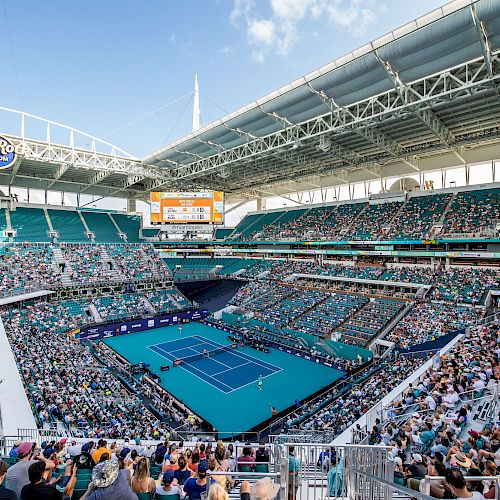 A packed stadium hosts a tennis match on a bright day, with spectators filling the stands around the central blue court.