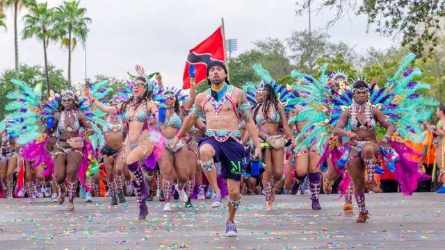 A group of dancers in colorful, feathered costumes perform energetically in a parade, with a red flag and palm trees in the background.