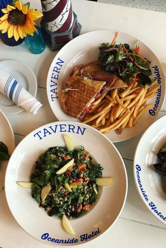 Plates of food including salads, sandwiches, fries, and a drink on a table labeled "Tavern Oceanside Hotel" with a sunflower centerpiece.