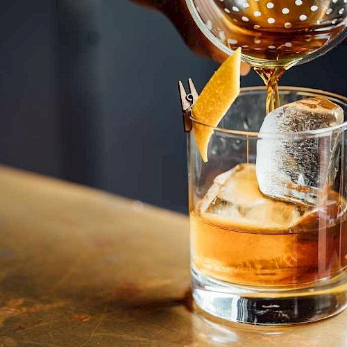 A classic cocktail being poured into a glass with ice, garnished with a twist of citrus peel clipped to the rim.