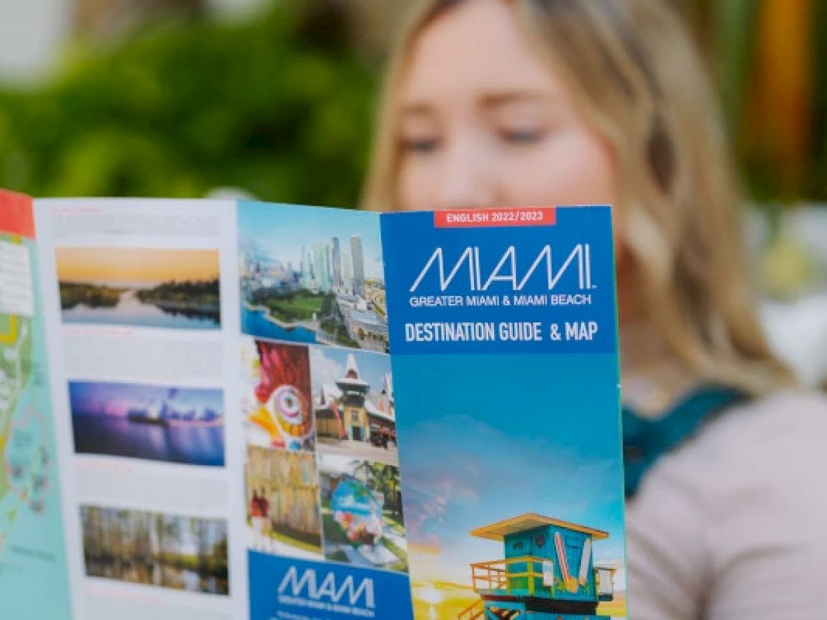 A person is reading a Miami travel guide, featuring images of beaches and attractions, titled "Destination Guide & Map" for 2022/2023.