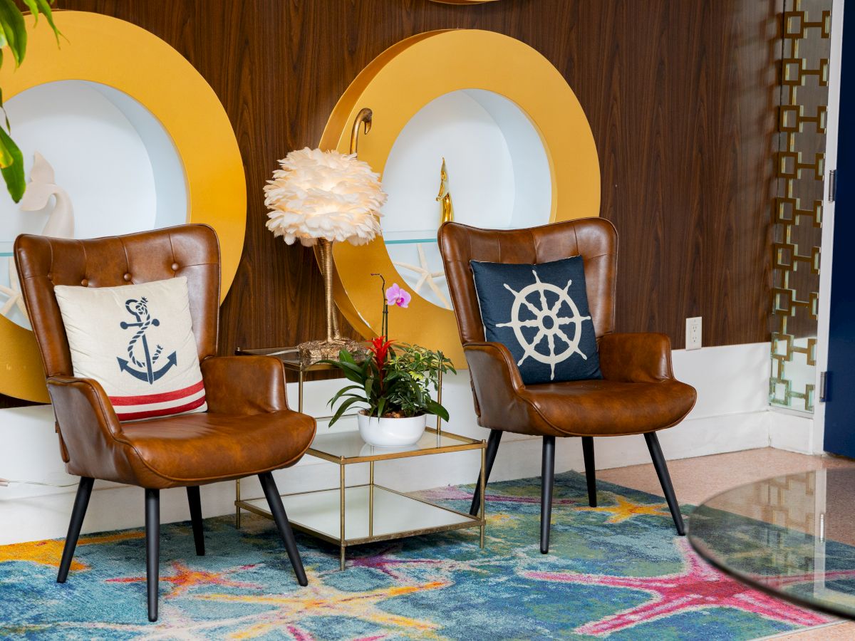 A stylish room with two brown chairs, nautical-themed pillows, a decorative lamp, plants, and colorful rug, set against wood paneling.