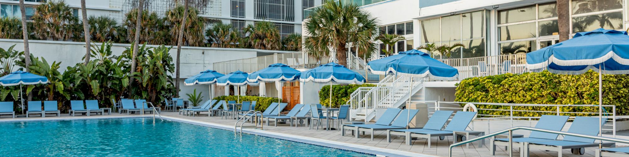 A luxurious outdoor pool area with blue sun loungers and umbrellas, surrounded by palm trees and modern buildings.