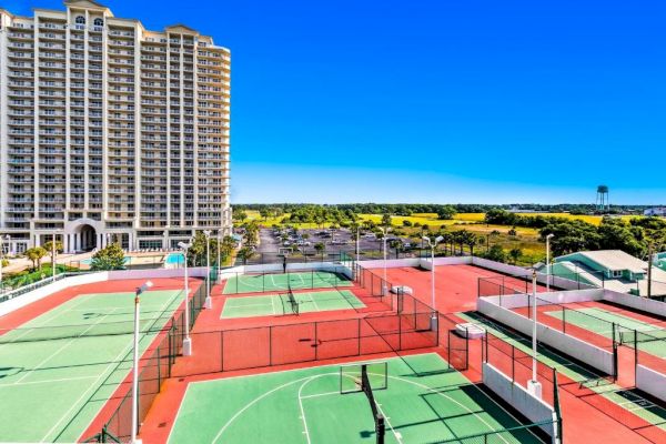 A residential high-rise building is beside tennis courts with red and green surfaces, surrounded by greenery under a clear blue sky.