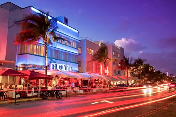 A vibrant street scene at night with neon-lit hotels, palm trees, and light trails from passing vehicles.