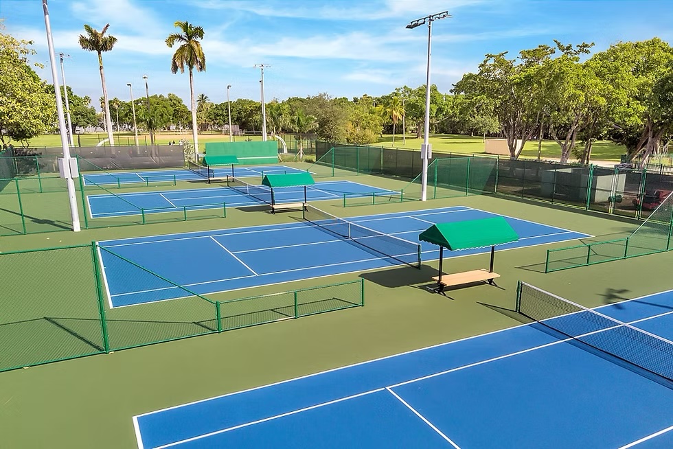 An aerial view of a tennis court with blue surfaces and green fencing, surrounded by palm trees and a clear blue sky.