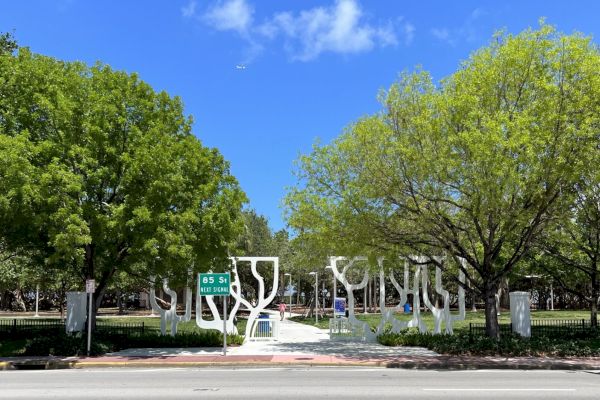 A park entrance with artistic white structures and green trees under a clear blue sky. A street sign indicates the next signal: 85 St.