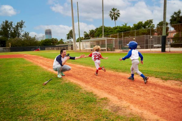 Two kids in baseball uniforms run toward a crouching adult on a baseball field, with one kid high-fiving the adult.