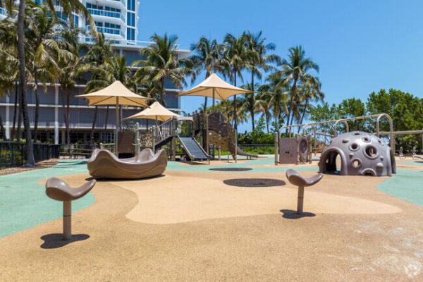 A playground with slides, climbing structures, and seats under palm trees, with a modern building in the background.