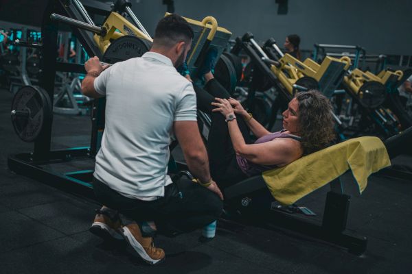 A person assists another using a leg press machine in a gym, with weights and equipment in the background.