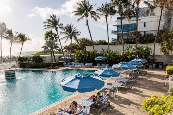 A tropical poolside scene with blue umbrellas, sun loungers, palm trees, and a nearby building.
