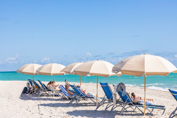 Beach scene with blue lounge chairs, beige umbrellas, and people relaxing on white sand by turquoise waters under a clear blue sky.