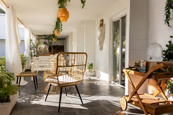 A cozy patio with wicker chairs, hanging plants, and warm lighting. There's a small cart and sliding glass door, creating a serene space.