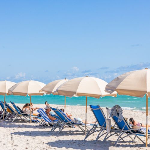 Beach scene with blue sky, parasols, lounge chairs, and people relaxing by the ocean.