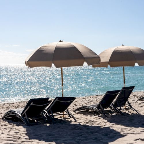 Beach scene with sandy shore, ocean view, two beige umbrellas, and several empty lounge chairs under a clear blue sky.