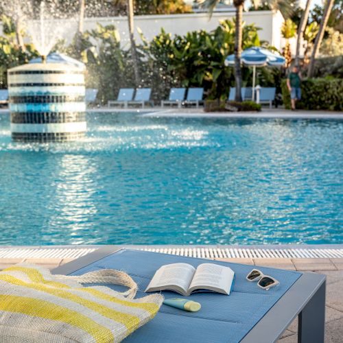 A poolside scene with a lounge chair, book, sunglasses, sunscreen, and striped bag; a fountain and palm trees in the background.