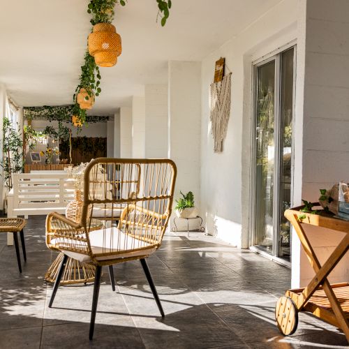 A cozy patio with wicker chairs, plants, a swing, lanterns, and a wooden cart under soft lighting in a serene setting.
