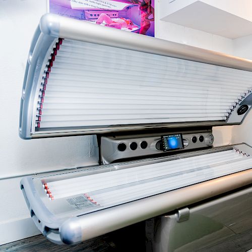A tanning bed in a room with a white chair and towels, under a wall light.