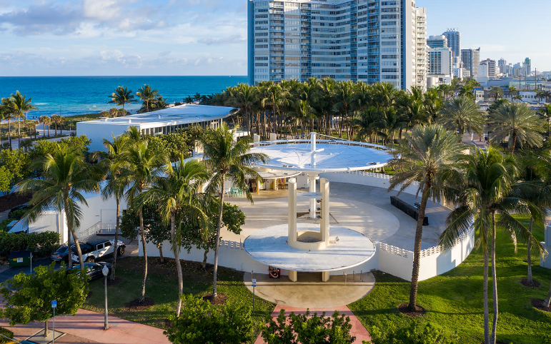 Aerial view of a white building with a curved roof surrounded by palm trees and a city skyline in the background.
